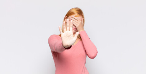 red head pretty woman covering face with hand and putting other hand up front to stop camera,...