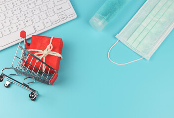  flat lay of red gift box in shopping cart, computer keyboard, medical mask and alcohol sanitizer gel on blue background with copy space. Covid19 prevention, new normal and online shopping.