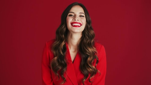 A friendly woman in a red blazer is doing hello gesture standing isolated over red background in the studio