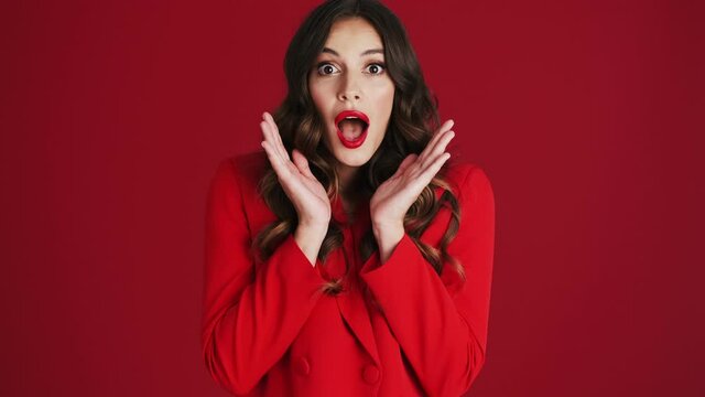 A surprised woman in a red blazer is opening her mouth standing isolated over red background in the studio