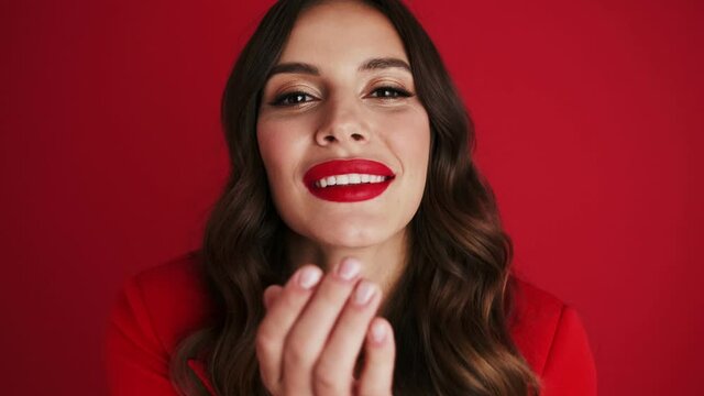 Close-up view of a smiling woman in a red blazer is sending an air kiss standing isolated over red background in the studio