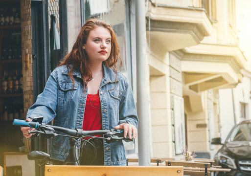 Red curled long hair caucasian teen girl on the city street walking with bicycle fashion portrait. Natural people beauty urban life concept image.