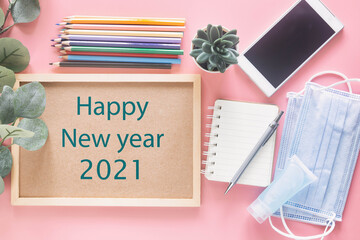 Happy New Year 2021 on wooden letter board with stationery, smartphone, mask and hand sanitizer on pastel pink background in top view flat lay. Concept to present new normal behavior in new year.