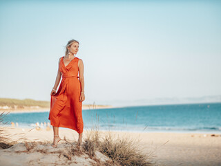 Pretty Blond Girl wearing a long red dress on the beach, playing with sand and looking towards the ocean