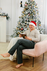 Portrait of a man with a book in a chair in the New Year's interior. Sports man in a Santa hat against the background of a Christmas tree. Christmas and New Year concept.