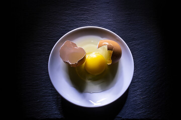 broken egg with shells on saucer over dark stone countertop. egg white yolk and egg shells