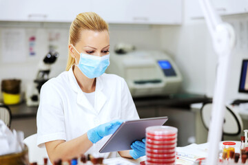 Innovative experienced female lab assistant in white sterile uniform with surgical mask and rubber gloves on sitting in laboratory and using tablet to enter test results.