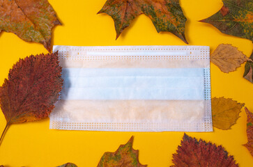 Creative composition of autumn leaves and a medical mask on a yellow background. Concept of the second wave of coronavirus infection. Postcard. Top view, space for advertising text