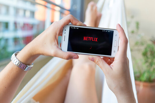 ROSARIO, ARGENTINA - JANUARY 19, 2020: Young woman in a hammock  opening Netflix  in her smartphone. Millennial girl resting with her cell phone in her hands and the Netflix logo in the screen.