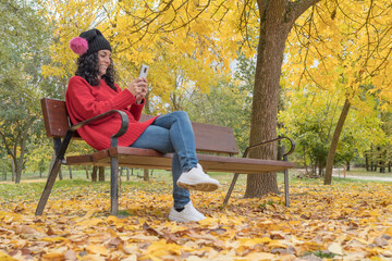 young woman in a woolen hat sitting on a wooden bench in a park laughing while chatting with her mobile. background of colored trees and fallen leaves