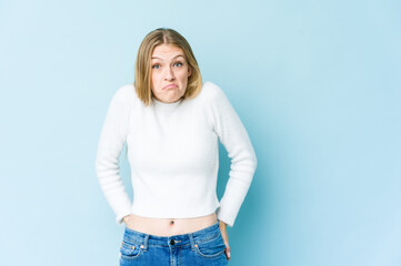 Young blonde woman isolated on blue background shrugs shoulders and open eyes confused.