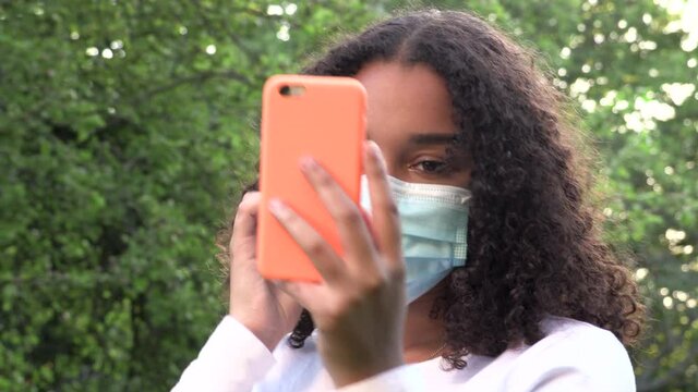 Beautiful mixed race African American girl teenager young woman wearing a face mask during COVID-19 Coronavirus pandemic using her smartphone or cell phone for social media in a park