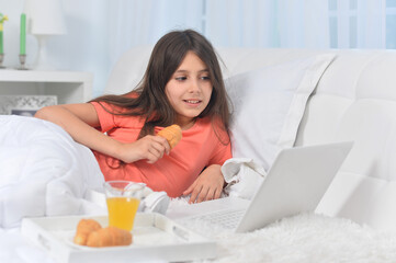 Obraz na płótnie Canvas Beautiful girl lying on soft beige bed with laptop and having breakfast