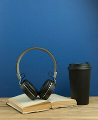 Black paper cup of coffee with headphones on wood desk with copy space. Photo concept of branding , cafe, leisure, reading, refreshment, relaxation.