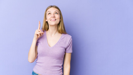 Young blonde woman isolated on purple background indicates with both fore fingers up showing a...