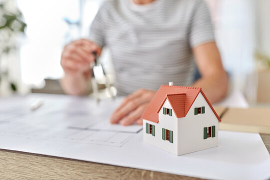 architecture, building, construction and real estate concept - living house model on table and male architect with compass measuring blueprint at home office
