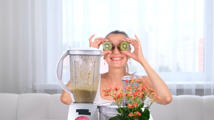 happy young woman preparing delicious nutritious smoothie in blender. funny female making eyes from kiwi.