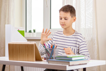 Kid with tablet computer sitting at table with books and having video call, virtual online leasson at home