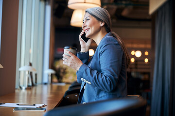 Cheerful businesswoman talking on cellphone and drinking coffee