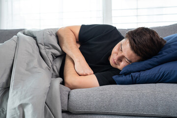 Fototapeta premium Sick and unwell Asian men holding his head, having a strong headache and sleeping on sofa at home wrapped in grey blanket
