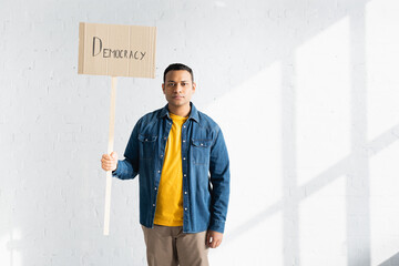 serious indian man holding placard with democracy inscription against white brick wall