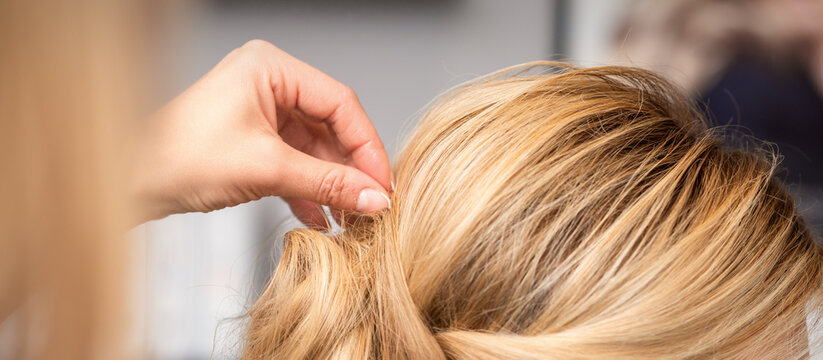 Close up back view of hand of female hairdresser makes hairstyle of young blonde woman in beauty salon