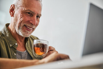 Smiley senior citizen having tea and checking his laptop