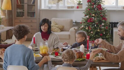 PAN shot of big family sitting at dinner table on Christmas day and holding hands while elderly man with grey hair and beard saying prayer before eating
