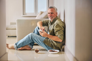 Joyful elderly man with his smartphone having tea at home