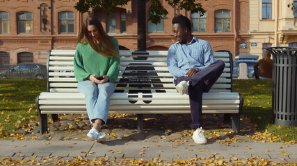 Young diverse people keeping social distancing sitting at bench outdoor