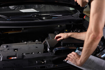 handsome caucasian guy is making notes while repairing hood of car, check all missing attributes inside of automobile