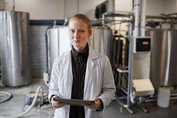Waist up portrait of young woman wearing lab coat posing against machines while working at dairy factory, copy space