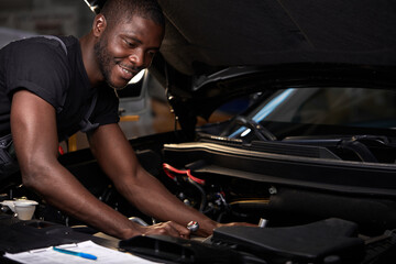 Fototapeta na wymiar african professional auto service technician in uniform standing near car hood repairing and using check list for car inspect, indoors