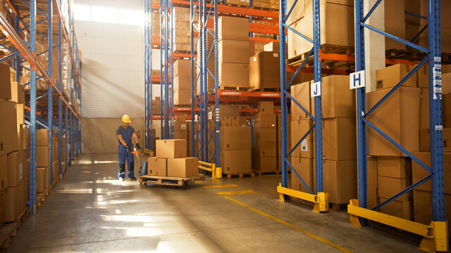 Worker Moves Cardboard Boxes Using Manual Pallet Truck, Walking Between Rows Of Shelves With Goods In Retail Warehouse. People Work In Product Distribution Logistics Center. Side View