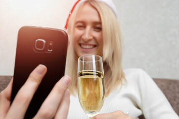 Young beautiful blonde woman in a Santa hat in a white sweater with a glass of champagne smiles and communicates via mobile phone close up. Concept of Christmas, New year, online communication.