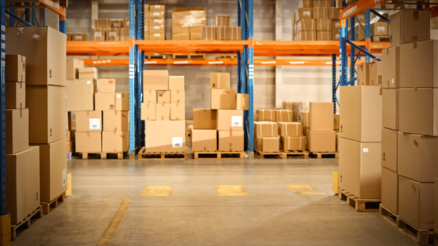 Gigantic Sunny Retail Warehouse Full Of Shelves With Goods In Cardboard Boxes. Logistics And Distribution Storehouse Center For Further Product Delivery Packages. Front Camera View