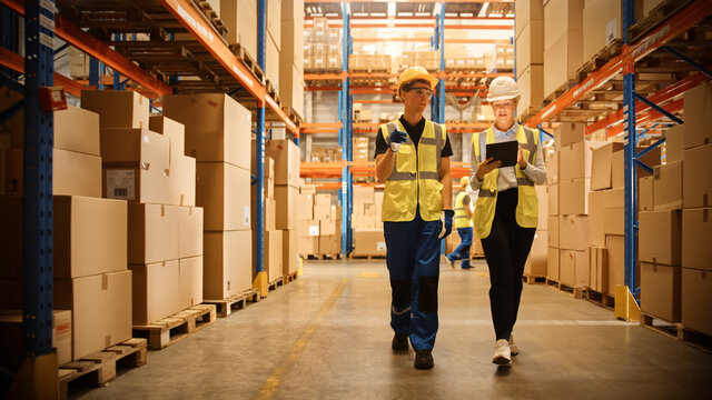 Retail Warehouse full of Shelves with Goods in Cardboard Boxes, Male Worker and Female Supervisor Holding Digital Tablet Discuss Product Delivery while Scanning Packages. Distribution Logistics Center
