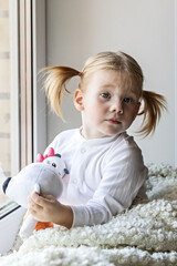 A little girl in white clothes plays with soft toys by the window. Self-isolation and quarantine content during the coronavirus pandemic in the world.