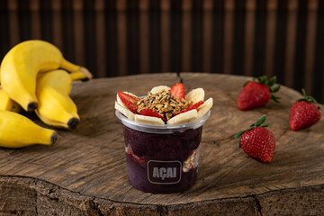 Delicious Brazilian Açaí Cream, in a plastic Cup With Strawberry, banana and granola Topping, in a rustic wooden background. Summer acai smoothie