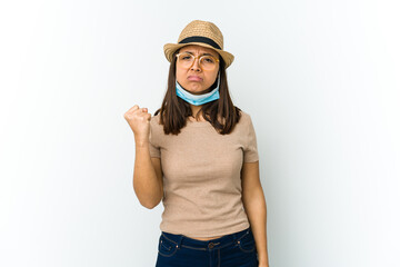 Young latin woman wearing hat and mask to protect from covid isolated on white background showing fist to camera, aggressive facial expression.