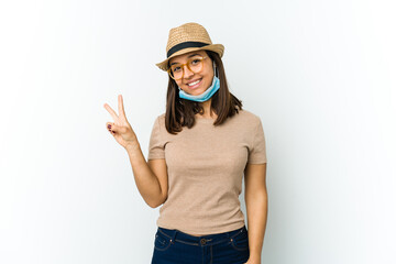Young latin woman wearing hat and mask to protect from covid isolated on white background joyful and carefree showing a peace symbol with fingers.