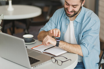 Cropped image of smiling young man sit alone at table in coffee shop cafe restaurant indoors working studying on laptop pc computer using smart watch on hand. Freelance mobile office business concept.