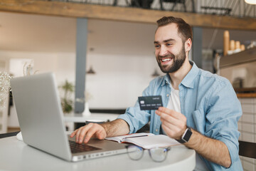 Excited cheerful young man sitting alone at table in coffee shop cafe restaurant indoors working or studying on laptop pc computer hold credit bank card. Freelance mobile office business concept.