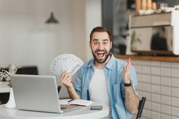 Excited man sit at table in coffee shop cafe restaurant indoors working studying on laptop computer hold fan of cash money in dollar banknotes spreading hand. Freelance mobile office business concept.