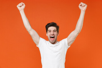Fototapeta premium Joyful screaming young man 20s in basic casual white blank empty t-shirt standing clenching fists doing winner gesture looking camera isolated on bright orange colour wall background, studio portrait.