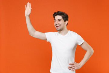 Laughing cheerful young man 20s wearing basic casual white blank empty t-shirt standing waving and greeting with hand as notices someone isolated on bright orange colour background, studio portrait.