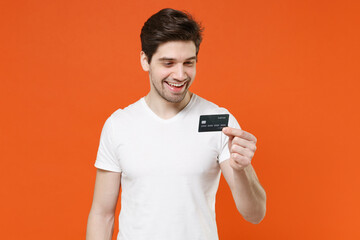 Smiling cheerful handsome attractive young man 20s wearing basic casual empty blank white t-shirt holding in hands credit bank card isolated on bright orange colour background studio portrait.