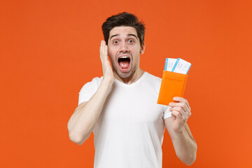 Shocked screaming young traveler tourist man in white t-shirt hold passport ticket put hand on cheek isolated on orange background. Passenger traveling on weekend getaway. Air flight journey concept.