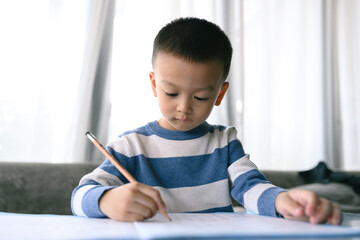 Asian Boy Doing homework with the intention. Child boy holding pencil writing, A boy drawing on white paper at the table, Elementary school and home schooling, Distance online Education concept.