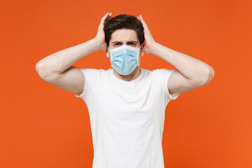 Worried young man wearing casual white t-shirt sterile face mask to safe from coronavirus virus covid-19 during pandemic quarantine put hands on head isolated on orange background studio portrait.